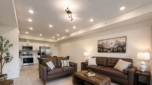 A modern living room with a brown leather sofa, a wooden coffee table, and a kitchen in the background. - New Pige Forge Condo | Mtn View - Walk Everywhere (Pigeon Forge)