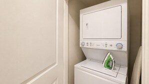A compact laundry room with a front-loading washer and dryer, a green iron, and a door leading to another room. - New Pige Forge Condo | Mtn View - Walk Everywhere (Pigeon Forge)