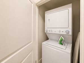 Una cambra de bugaderia petita amb rentadora i assecadora frontals, un ferró verd i una porta que dona a una altra habitació.