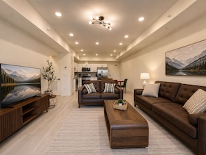 A modern living room with a flat-screen TV, leather sofas, a wooden coffee table, and a kitchen in the background. - New Pige Forge Condo | Walk Everywhere (Pigeon Forge)