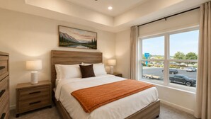 Quarto de hotel com uma cama grande, mesa de cabeceira com abajur, pintura na parede e janela com vista para o estacionamento.