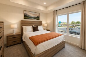 A hotel room with a large bed, a nightstand with a lamp, a painting on the wall, and a window with a view of the parking lot. - New Pige Forge Condo | Mtn View - Walk Everywhere (Pigeon Forge)