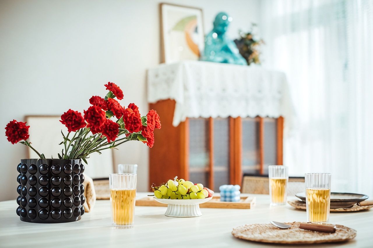 Una mesa de comedor con un jarrón de flores rojas, un cuenco de uvas verdes y vasos de cerveza.