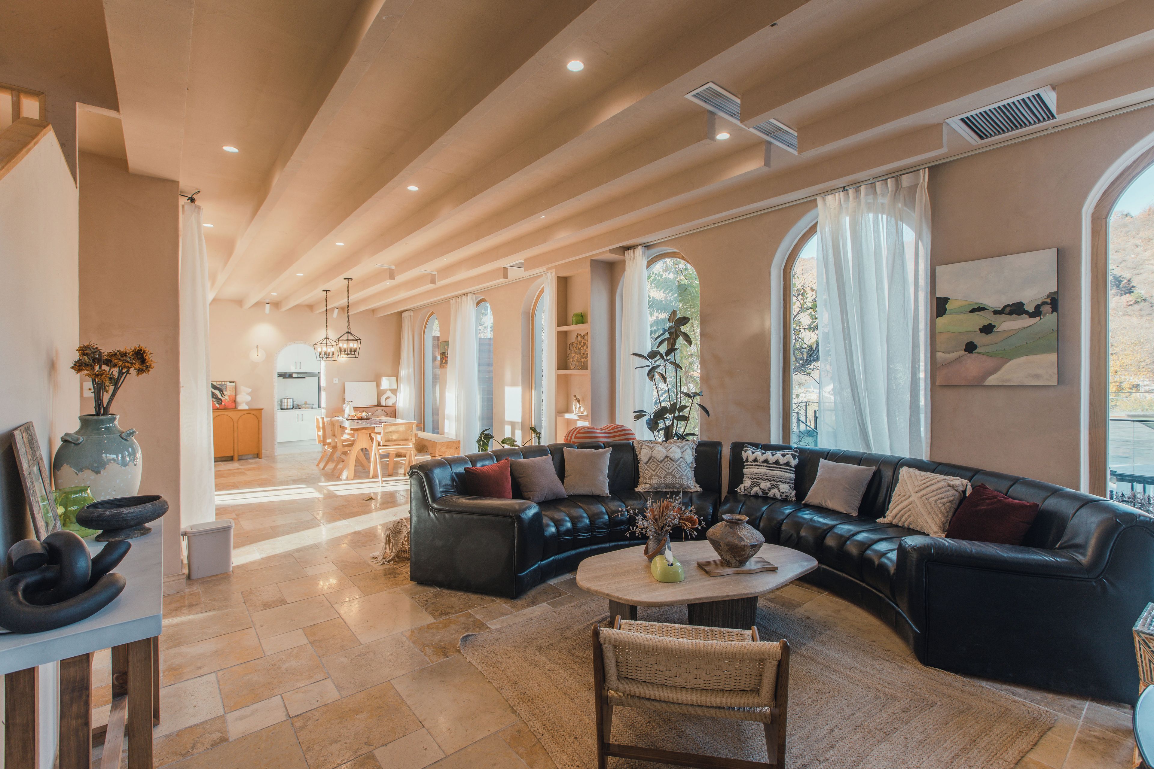 Amplia sala de estar con un sofá de cuero negro, un comedor con mesa de madera y ventanas grandes con cortinas.