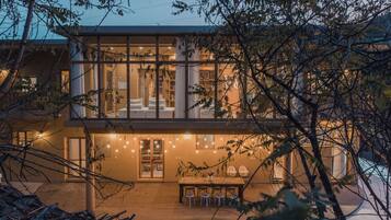 A modern two-story house with a balcony, a dining area with a table and chairs, and a view of the surrounding trees.