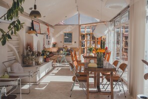 Un comedor amplio con techo alto, luz natural proveniente de ventanas grandes y diversas opciones de asientos, incluyendo una larga mesa de madera y sillas.
