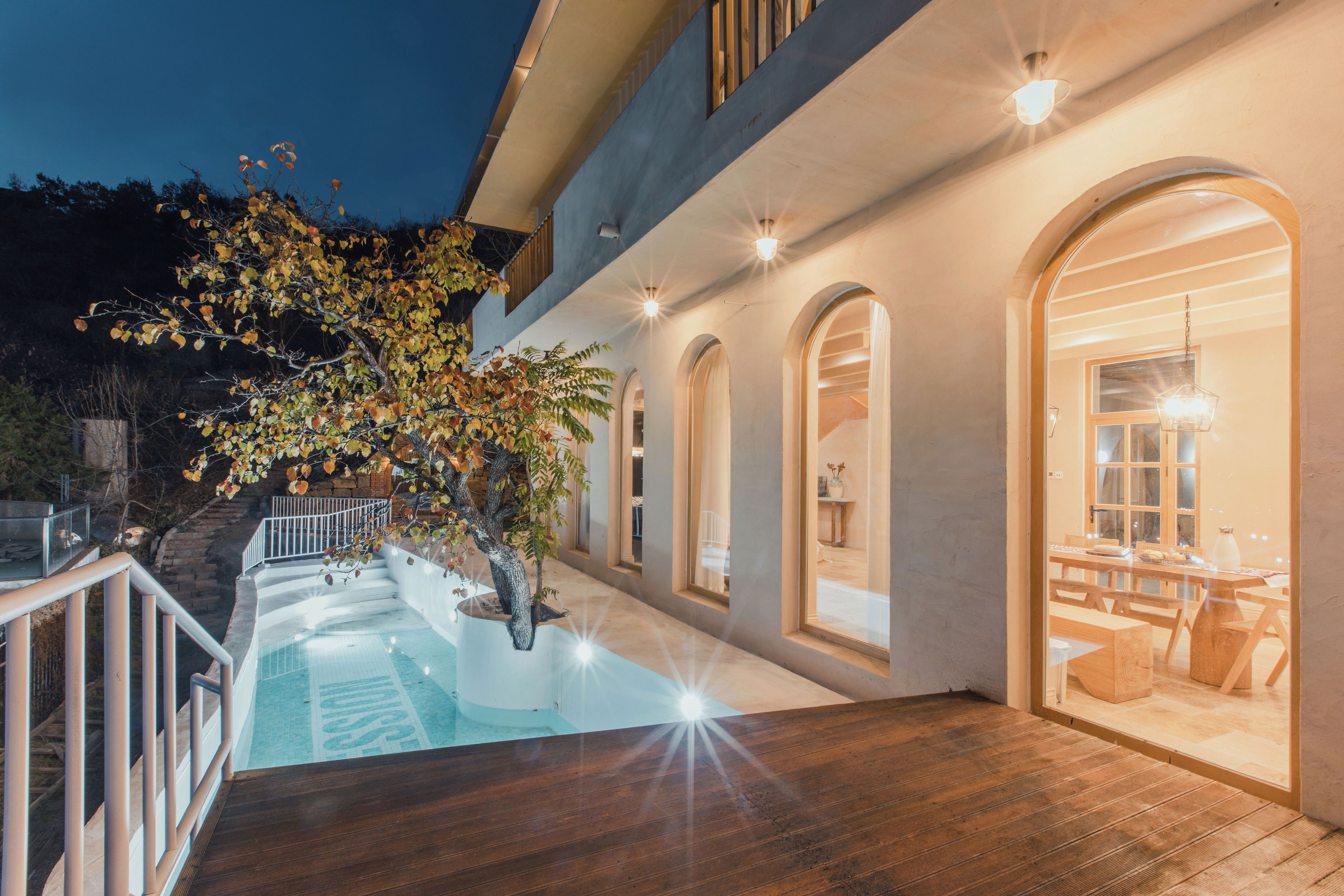 A swimming pool with a wooden deck, surrounded by a building with arched windows and a dining area visible through one of the windows.