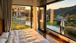A bedroom with a large bed, a glass balcony, and a view of a building and hills. - Qiran Rustic Resort Villa (Huairou)