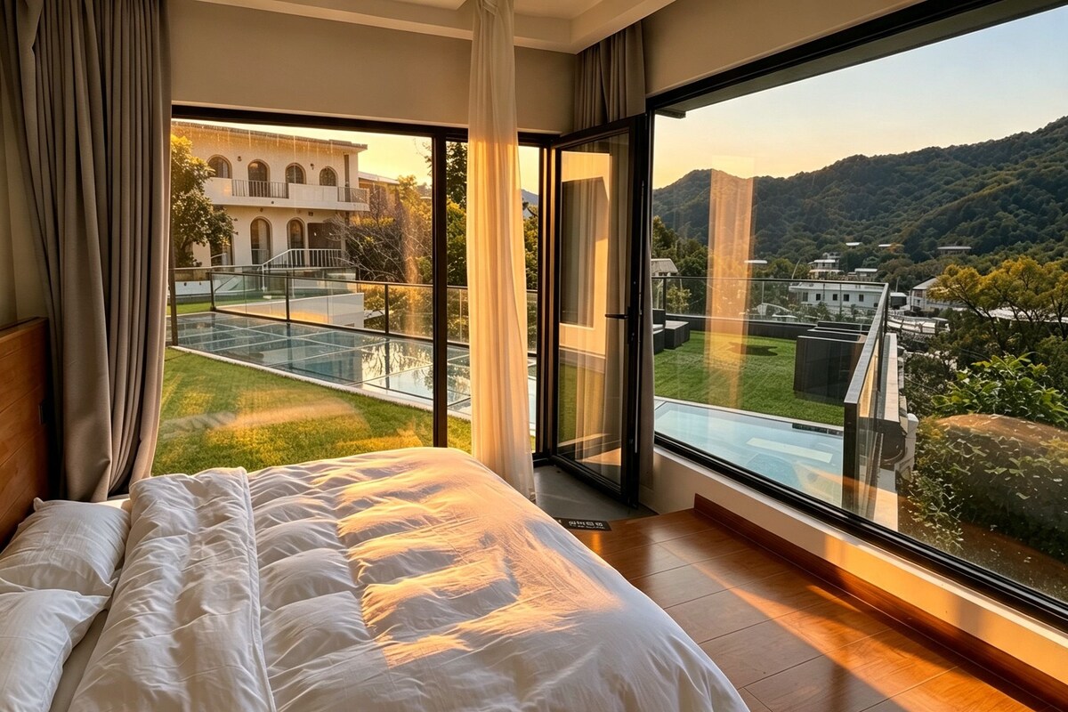 Un dormitorio con una cama grande, un balcón de vidrio y vistas a un edificio y colinas.