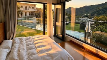 Un dormitorio con una cama grande, un balcón de vidrio y vistas a un edificio y colinas.