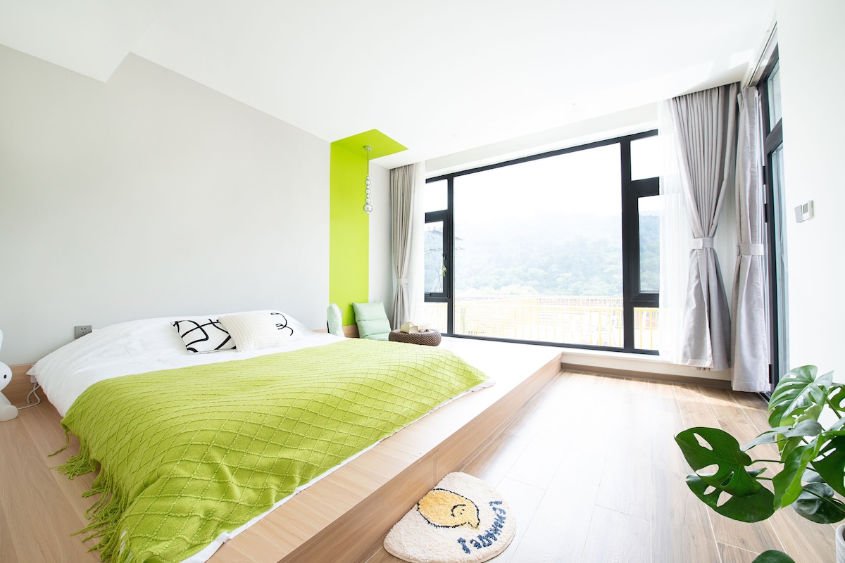 Un dormitorio moderno con un ventanal grande, una cama con ropa de cama verde y vistas a un paisaje urbano.