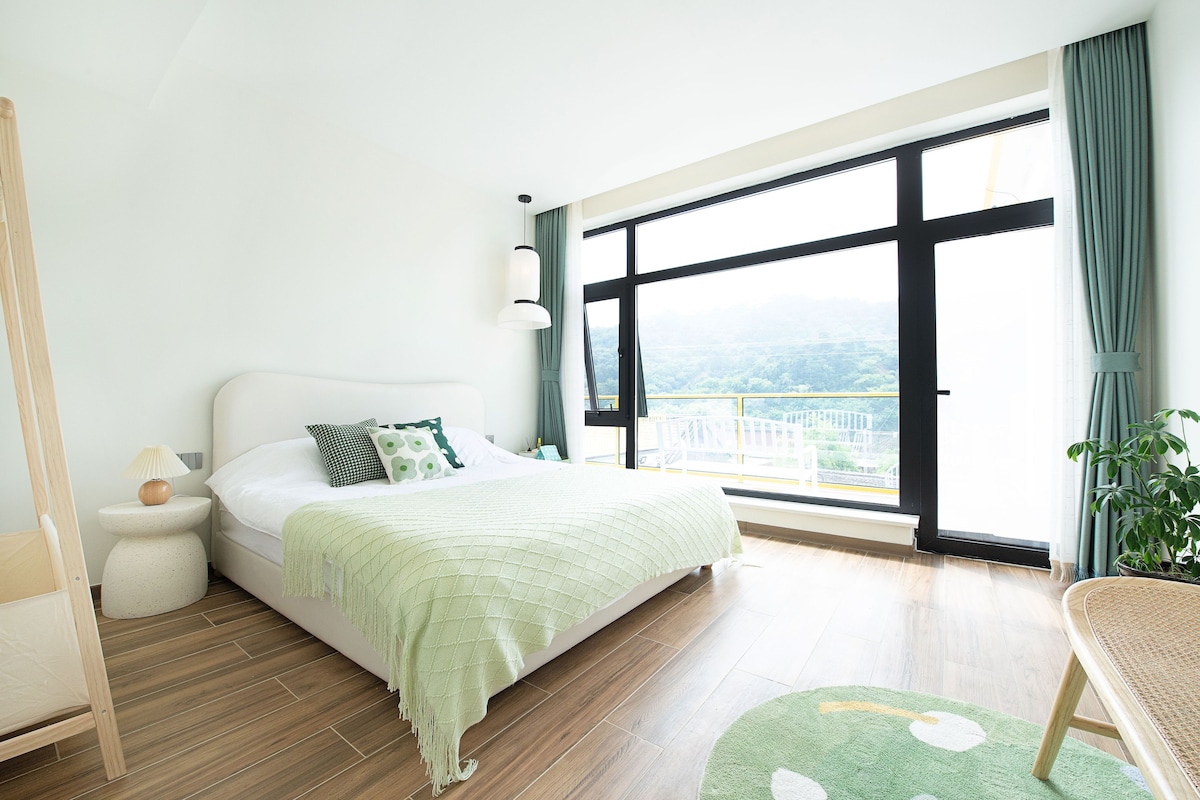 Un dormitorio con un ventanal grande, una cama con manta verde, una mesita de noche con lámpara y una planta en maceta.