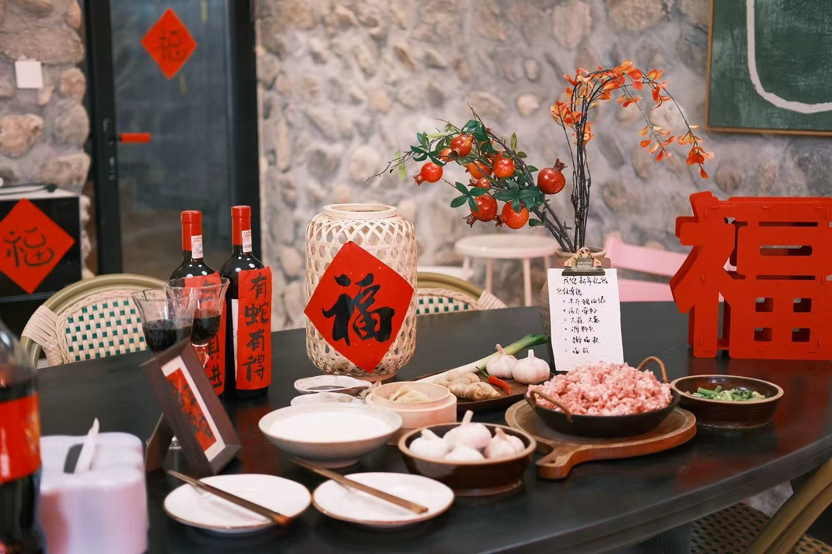 Una mesa dispuesta con diversos platos, incluyendo un tazón de dumplings, un plato de carne en rodajas y un jarrón con flores rojas. También hay botellas de vino y un cartel decorativo rojo con caracteres chinos.