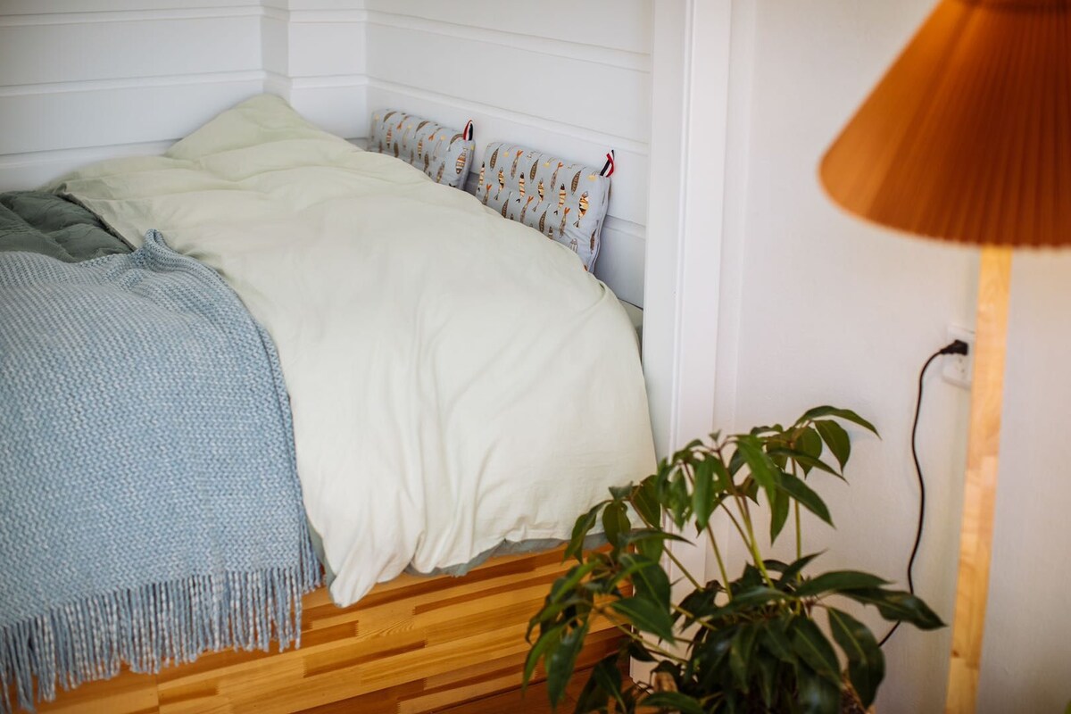 Una cama con edredón blanco, una manta gris y almohadas estampadas. Hay suelo de madera y una planta junto a la cama.