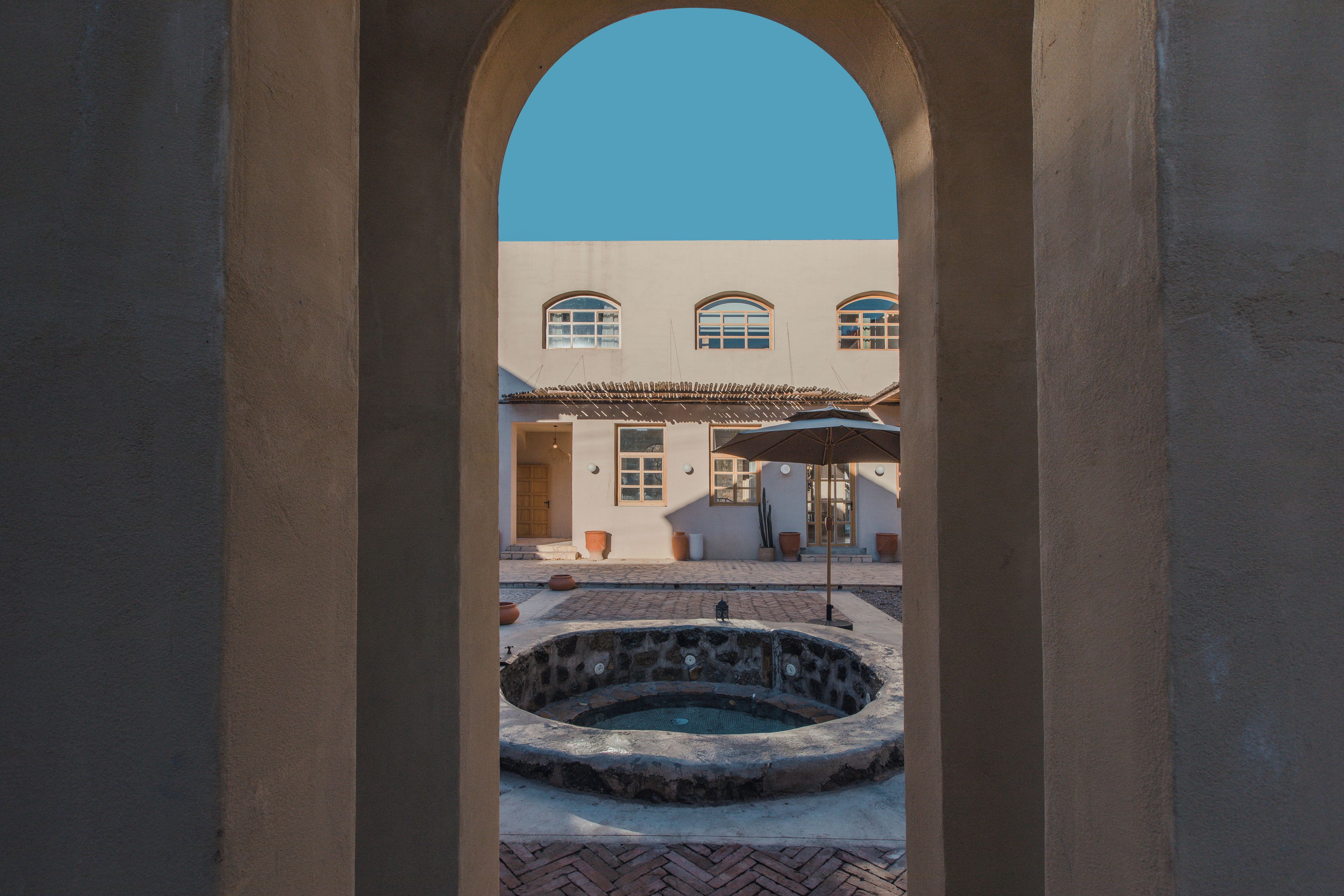 Uma porta em arco dá acesso a um pátio com uma fonte de pedra, chão de azulejos e um edifício com janelas em arco e telhado em azulejos.
