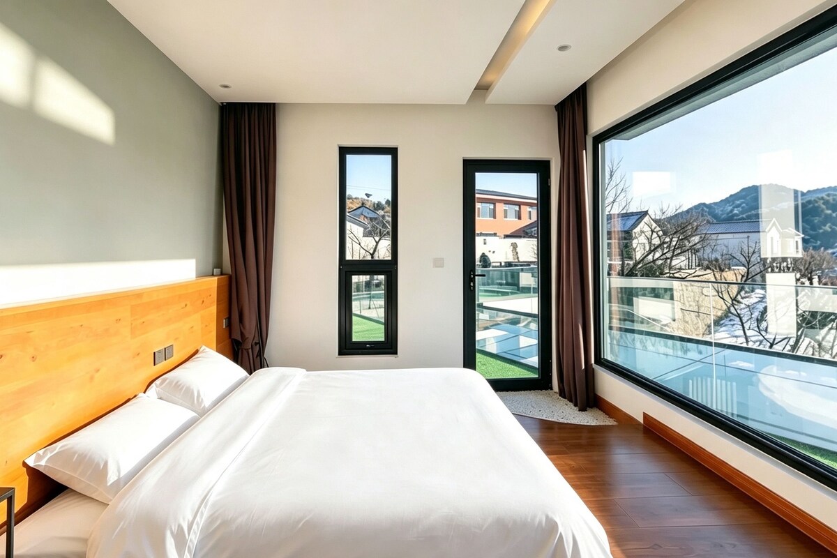 Un dormitorio moderno con una cama grande, cabecera de madera, vistas a un paisaje urbano a través de un ventanal y un balcón con barandilla de vidrio.