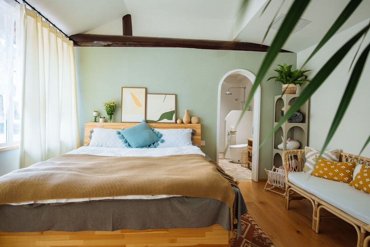 Un dormitorio con cama, mesita de noche, silla, estante con objetos decorativos y un acceso a un baño.
