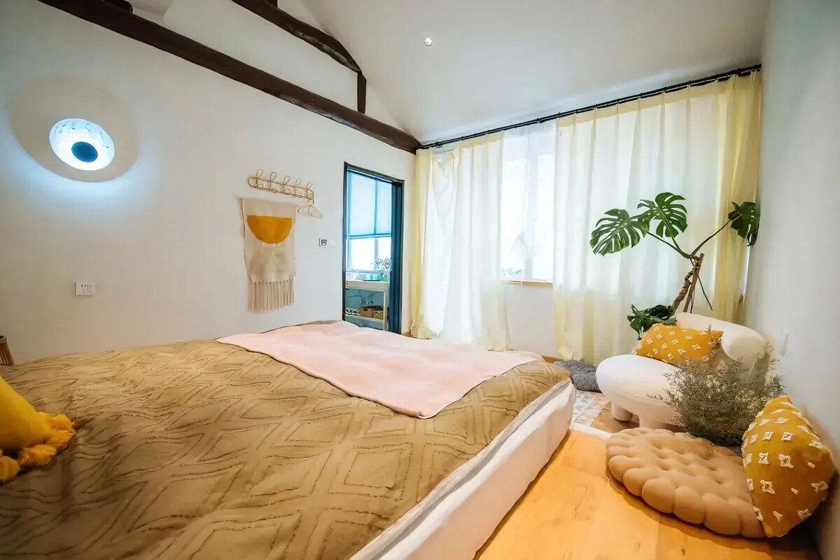 Un dormitorio con cama, mesita de noche, planta y ventana con cortinas.