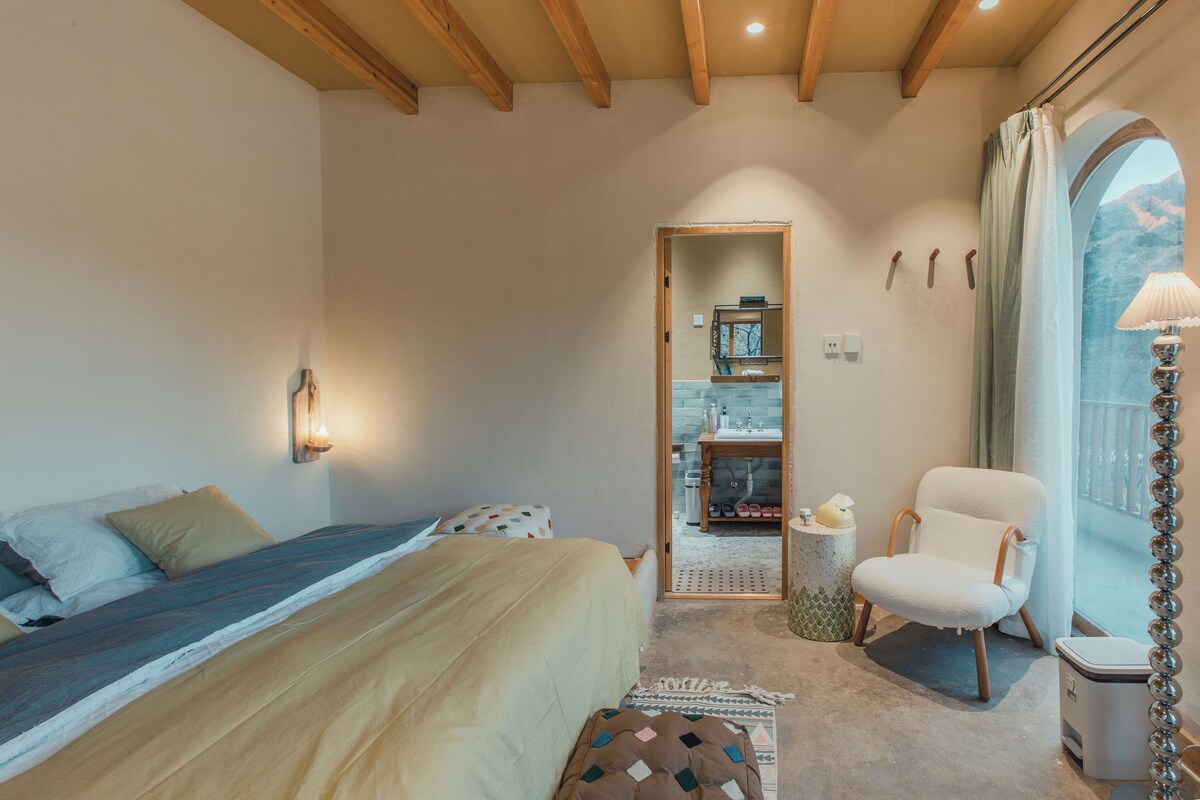 Un dormitorio con cama, una silla, un ventanal y un baño visible a través de una puerta abierta.