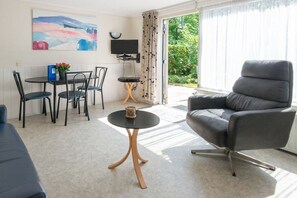 A modern living room with a sofa, a dining table with chairs, a TV mounted on the wall, and a view of greenery outside. - Holiday apartment with 1 bedroom (Zoutelande)