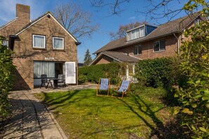 Een woonhuis met twee verdiepingen, een bakstenen gevel, een zichtbaar raam en een deur. Er is een terras met een verharde vloer, twee stoelen en een tafel.