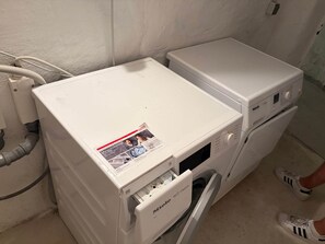 Eine weiße Waschmaschine mit Bedienfeld und kleinem Fenster steht in einer Ecke neben einer weißen Wand und einer Rohrleitung.