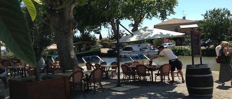 An outdoor dining area with tables and chairs, a large tree, and a boat in the background.