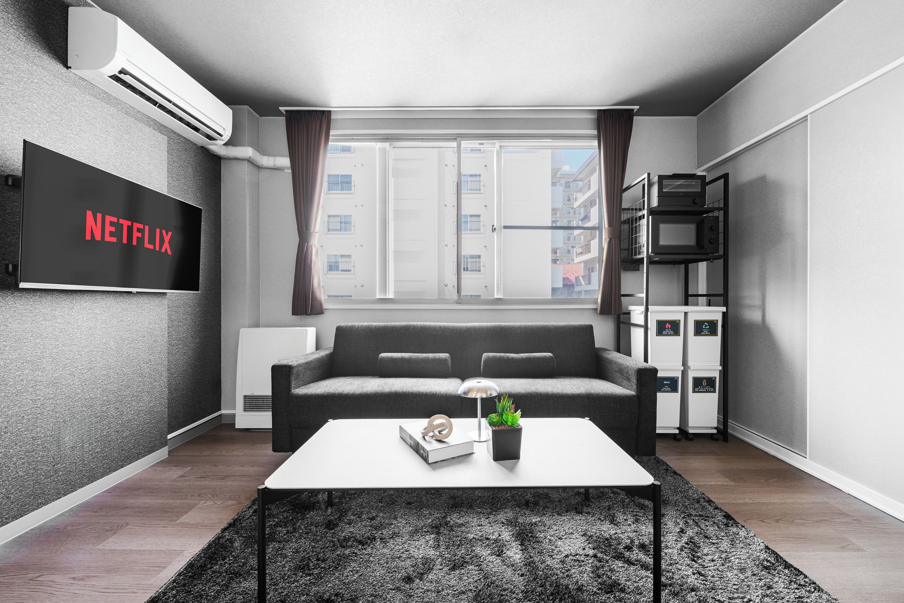 Un salón moderno con un sofá gris, una mesa de centro blanca con una planta, un televisor de pantalla plana en la pared y una ventana con cortinas.
