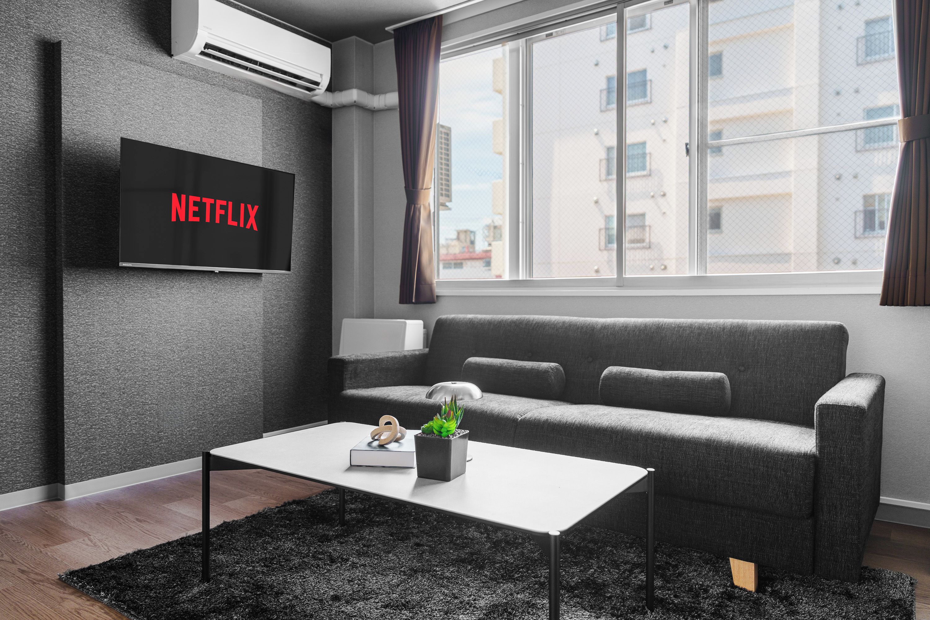 A modern living room with a grey sofa, a white coffee table, a flat-screen TV displaying 'NETFLIX', and a window with a view of buildings.
