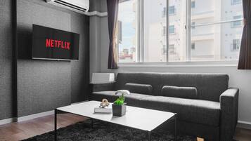 A modern living room with a grey sofa, a white coffee table, a flat-screen TV displaying 'NETFLIX', and a window with a view of buildings.