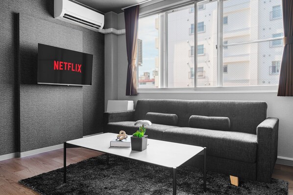 A modern living room with a grey sofa, a white coffee table, a flat-screen TV displaying 'NETFLIX', and a window with a view of buildings.