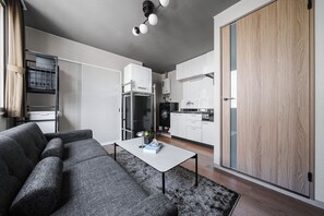 A modern apartment with a grey sofa, a small table with a book, a kitchen with white cabinets, and a wooden wardrobe.