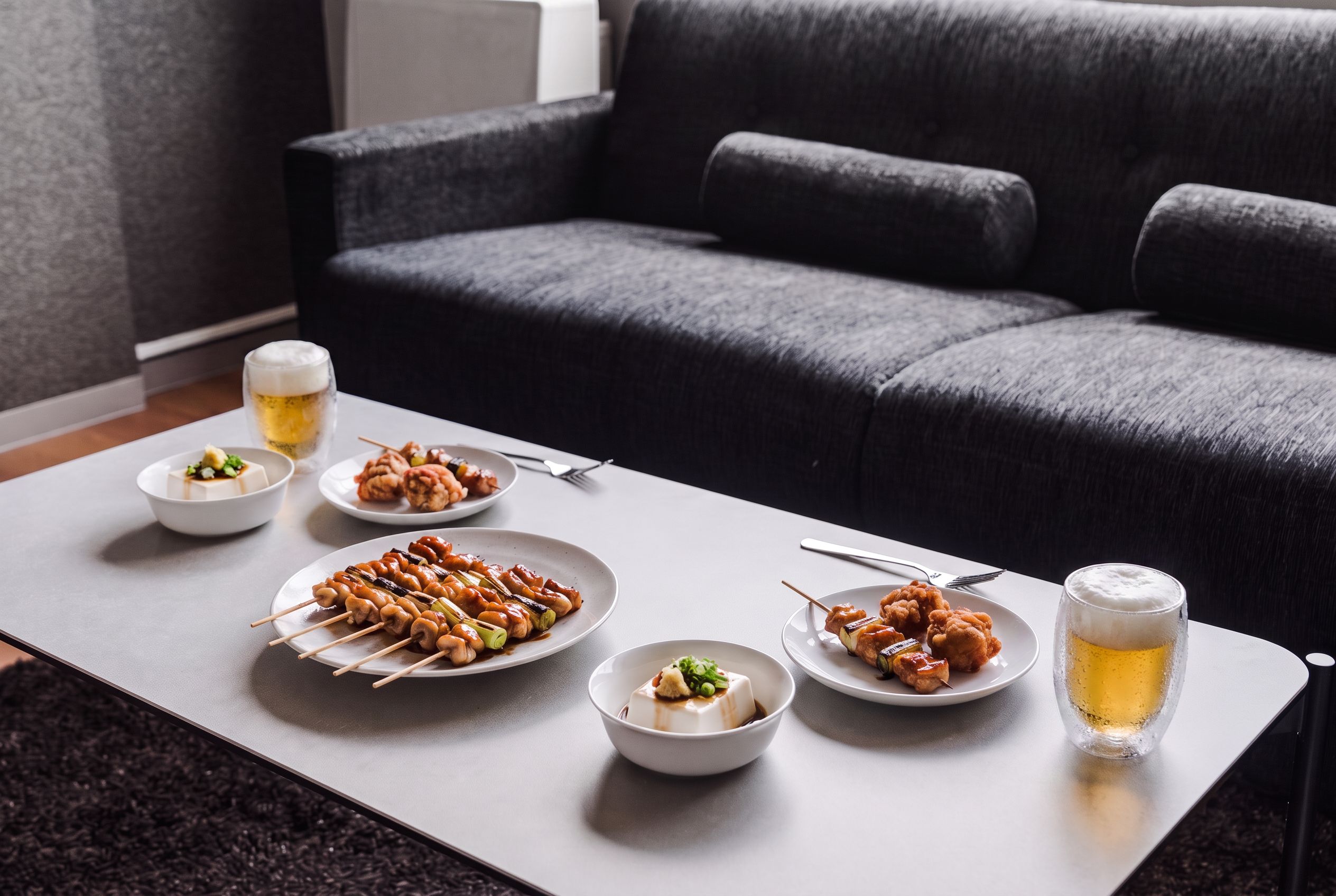 Uma mesa de jantar com pratos de comida, um copo de cerveja e um sofá ao fundo.