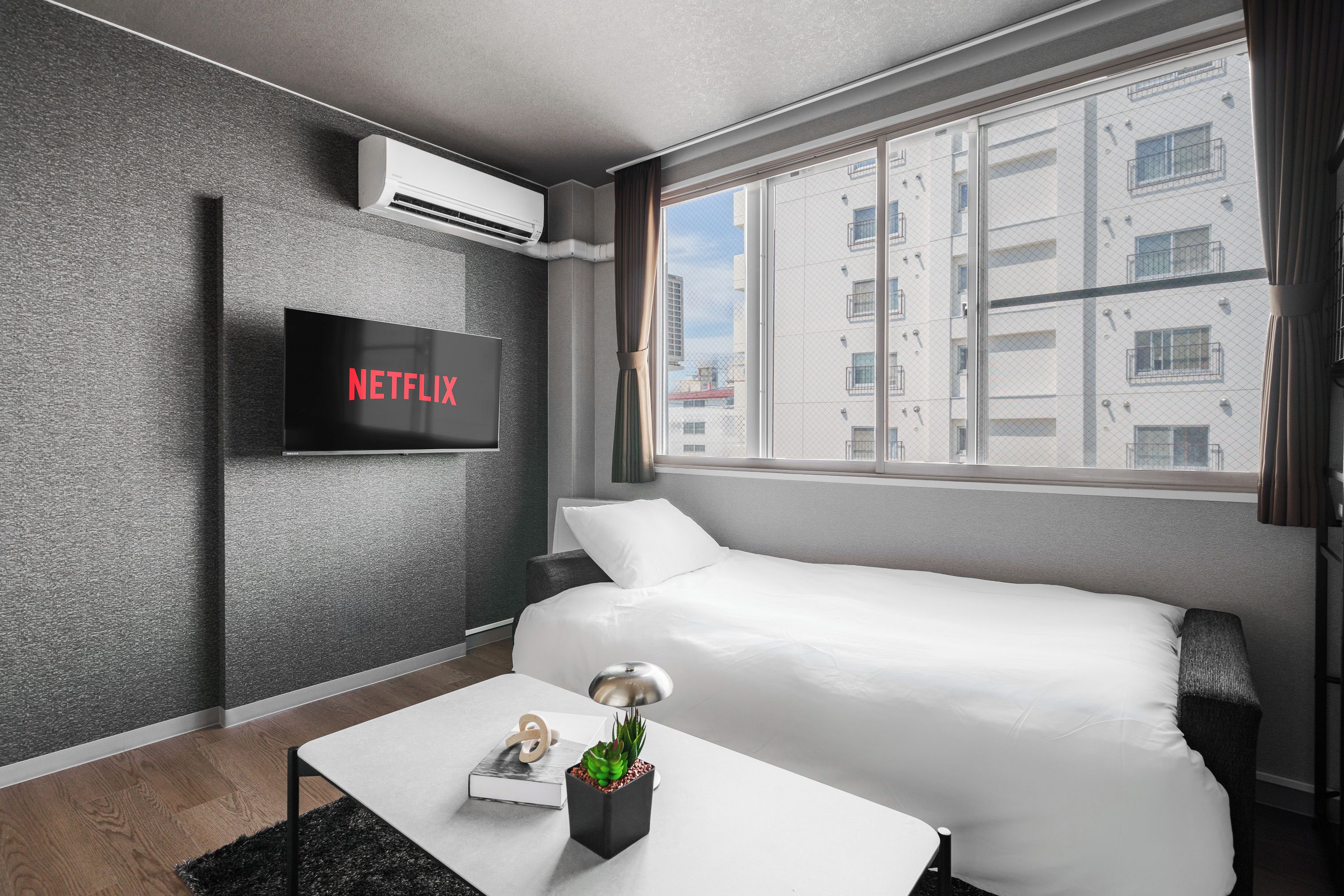 A modern hotel room with a bed, a flat-screen TV mounted on the wall, a small table with a lamp, and a window with a view of buildings.