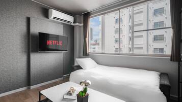 A modern hotel room with a bed, a flat-screen TV mounted on the wall, a small table with a lamp, and a window with a view of buildings.