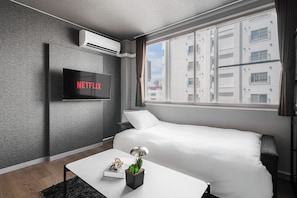 A modern hotel room with a bed, a flat-screen TV mounted on the wall, a small table with a lamp, and a window with a view of buildings.