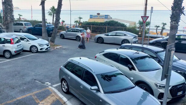 Un parking où sont garées plusieurs voitures, avec des palmiers et une vue sur la mer.