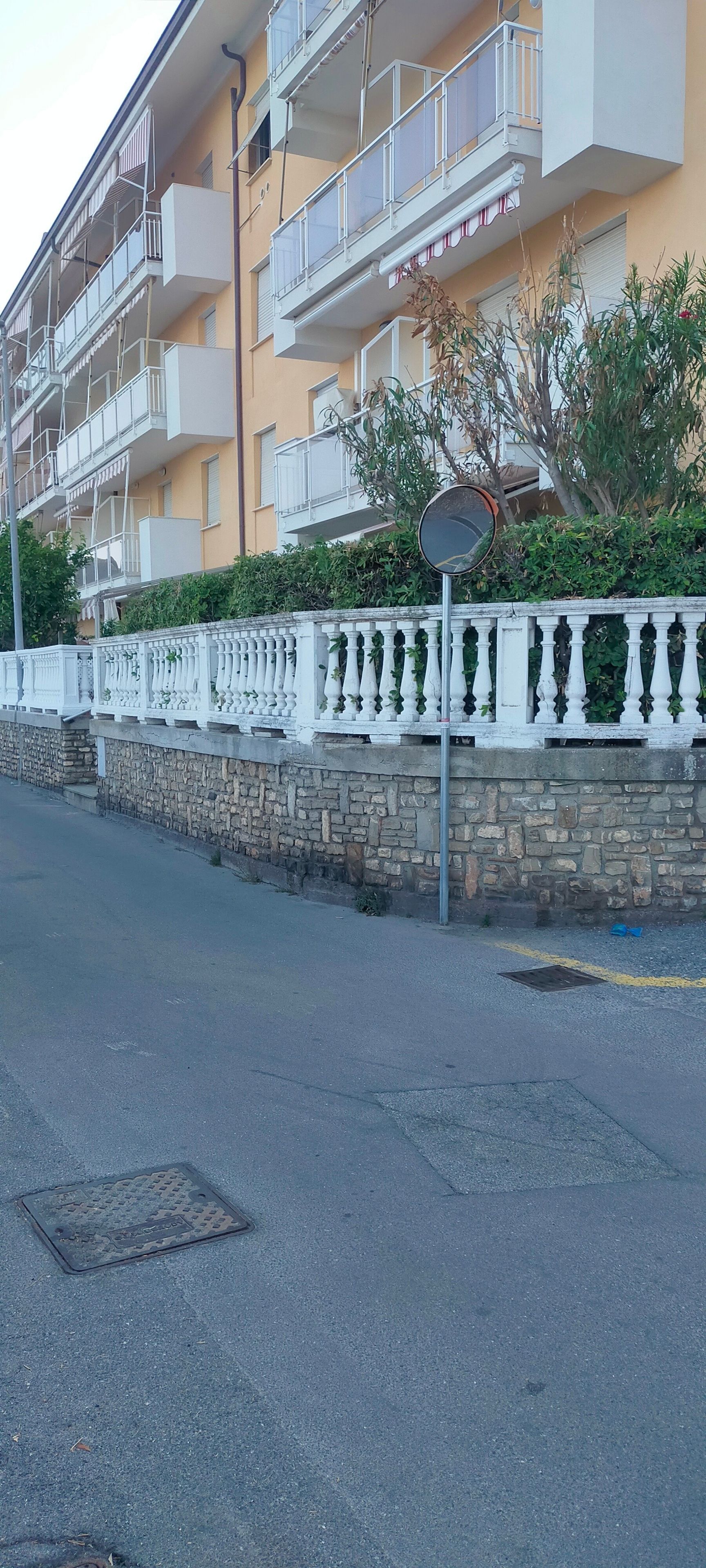 Un edificio residenziale a più piani con balconi, un muro in pietra con balaustra bianca e una strada di fronte.
