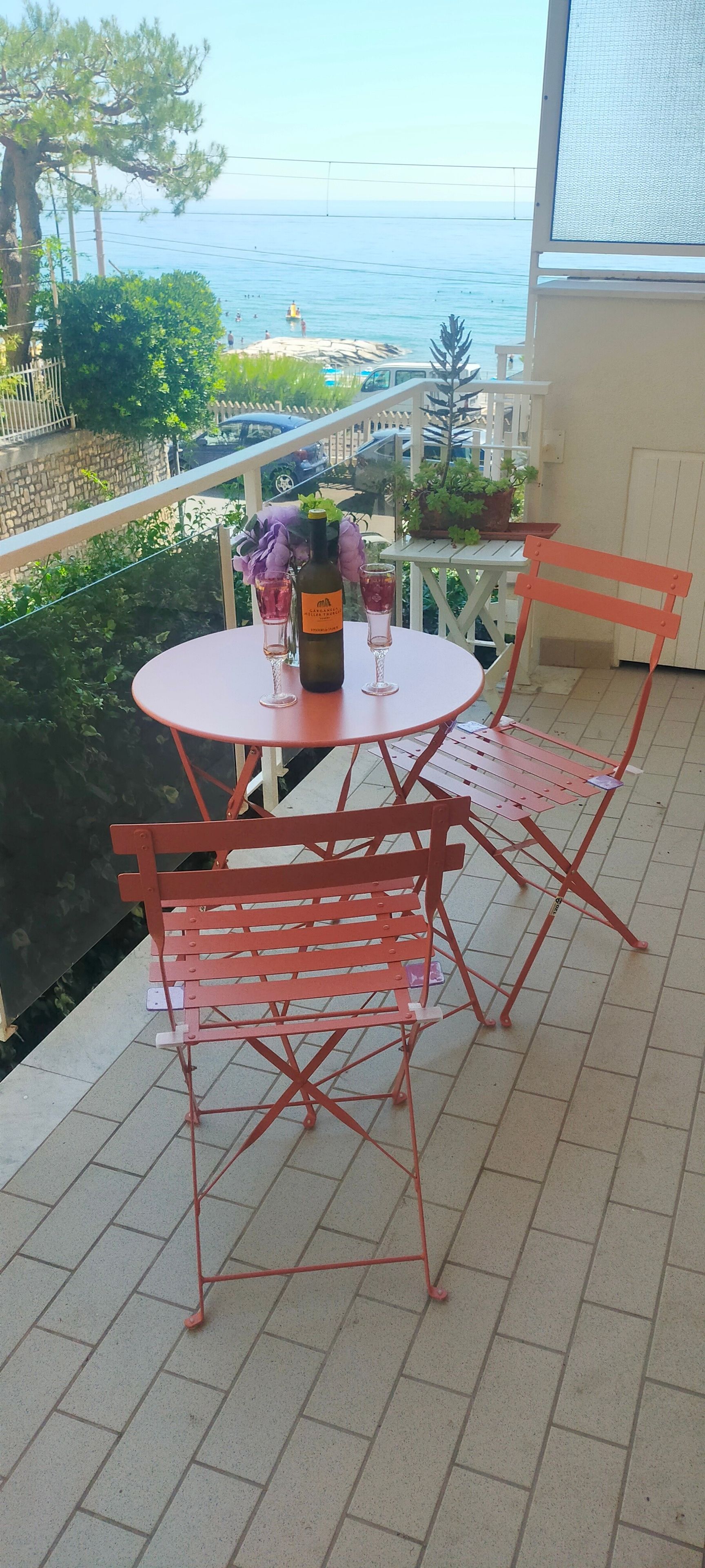 Een ronde tafel met vier stoelen, een fles en twee glazen op een balkon met uitzicht op zee.