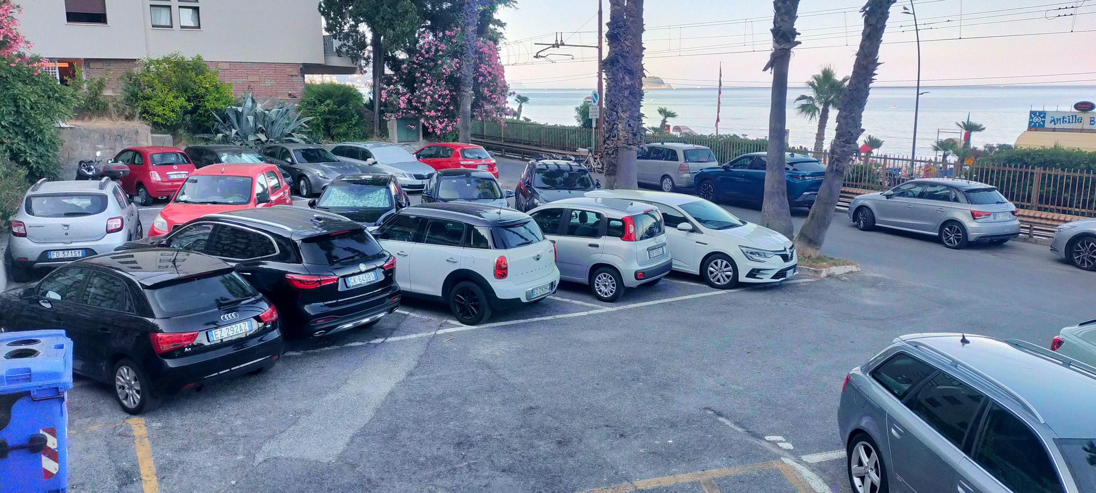 A parking lot with various cars parked, a building in the background, and a view of the sea.