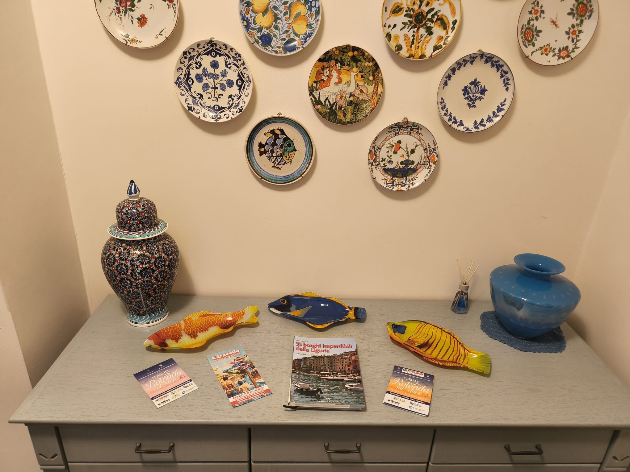 Een verzameling decoratieve borden, een keramieken vis, een blauwe vaas en enkele ansichtkaarten op een tafel.