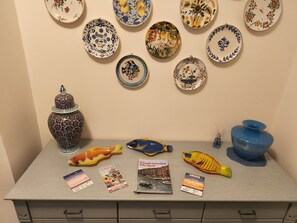 Esposizione di piatti decorativi, un pesce in ceramica, un vaso blu e alcune cartoline su un tavolo.