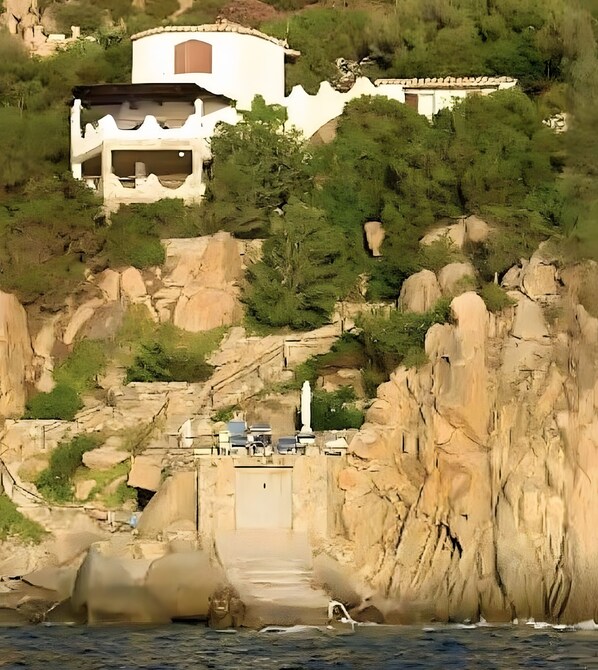 A coastal building with a stone facade, a staircase leading to a dock, and a statue in the foreground.