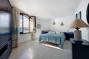 A bedroom with a bed, a desk, a lamp, a window with curtains, and a view of the sea.