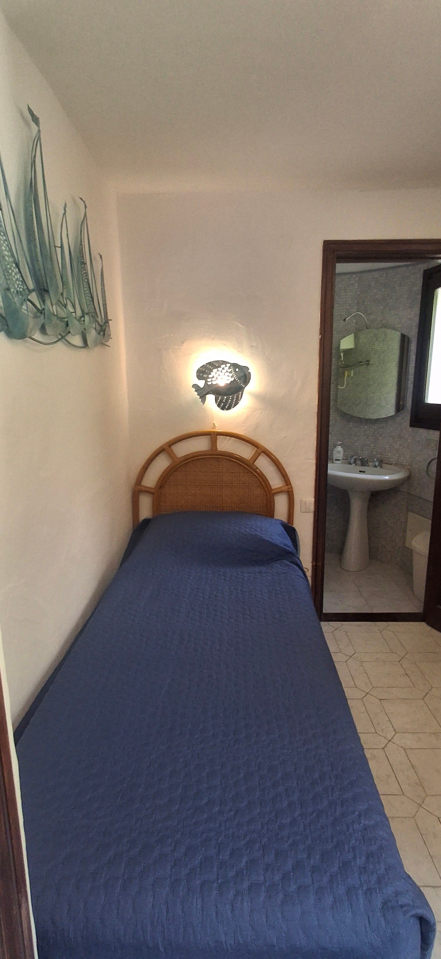 A small, single-bed room with a wooden headboard, a wall-mounted light fixture, and a bathroom visible through an open door.