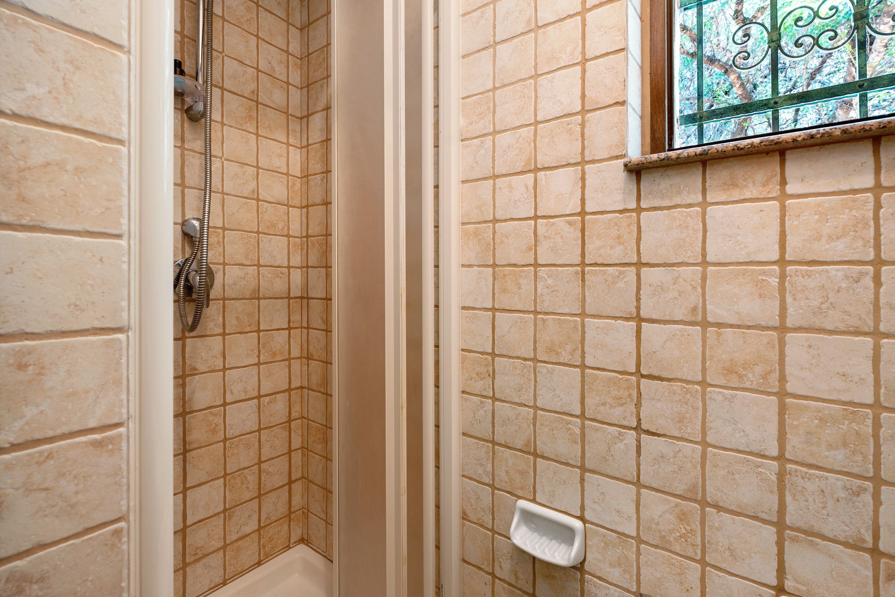 Un bagno con pareti piastrellate color crema, una doccia con porta in vetro e una piccola finestra con inferriate decorative.