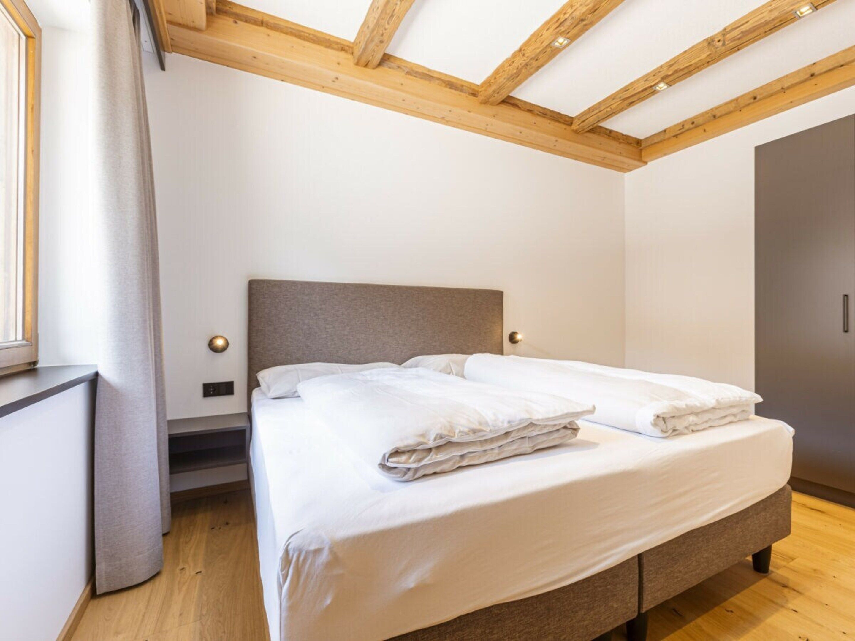 A bedroom with a bed, wooden ceiling beams, a window with curtains, and a wardrobe.