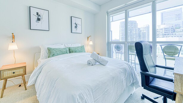 Een hotelkamer met een groot bed, twee kunstwerken aan de muur, een raam met uitzicht over de stad, een stoel en een nachtkastje met een lamp.