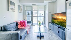 Een moderne woonkamer met een grijze bank, een glazen salontafel en een grote flatscreen televisie op een houten kast.