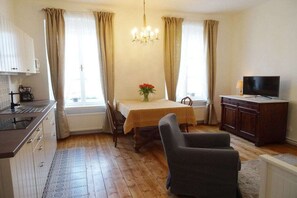 A room with a dining table, chairs, a television, a kitchen area, and a chandelier.
